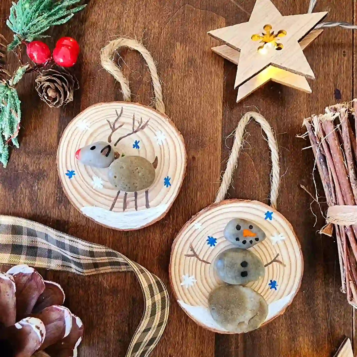 Two round wood slice ornaments with twine loops, one with a pebble reindeer with a red nose and one with a stacked pebble snowman, on a dark wooden table with pine cones and ribbon.