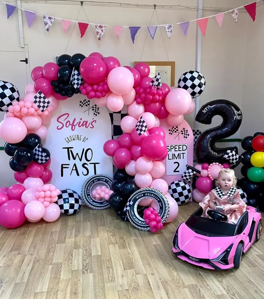 Hot pink, blush and black balloons around a white arch reading TWO FAST with a speed limit sign, checkered foil balls, tire balloons, a balloon traffic light and a toddler in a satin dress with a checkered collar in a pink ride-on car