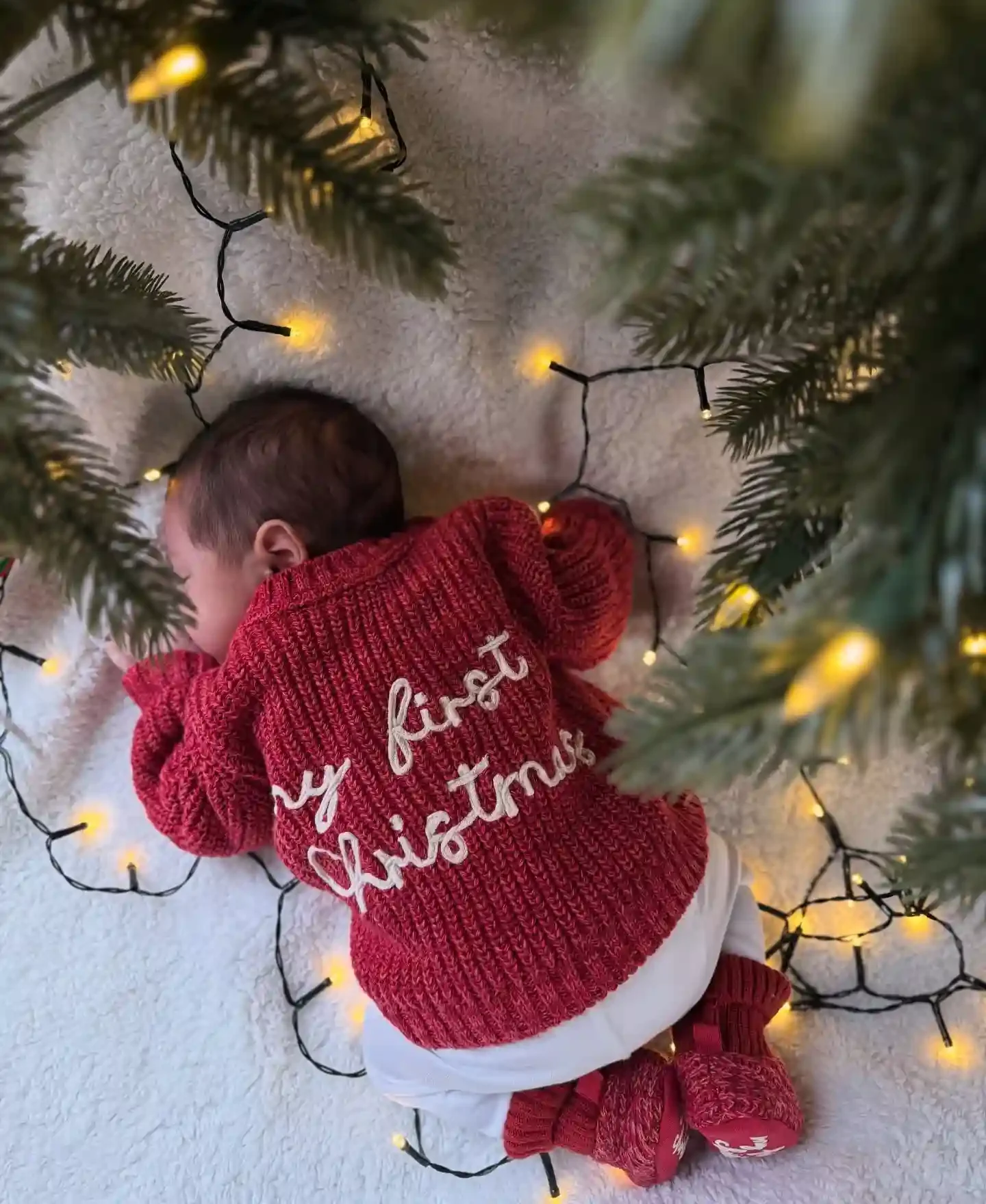Sleeping newborn on a white sherpa blanket in a chunky red cardigan stitched with my first Christmas, red knit booties and warm string lights
