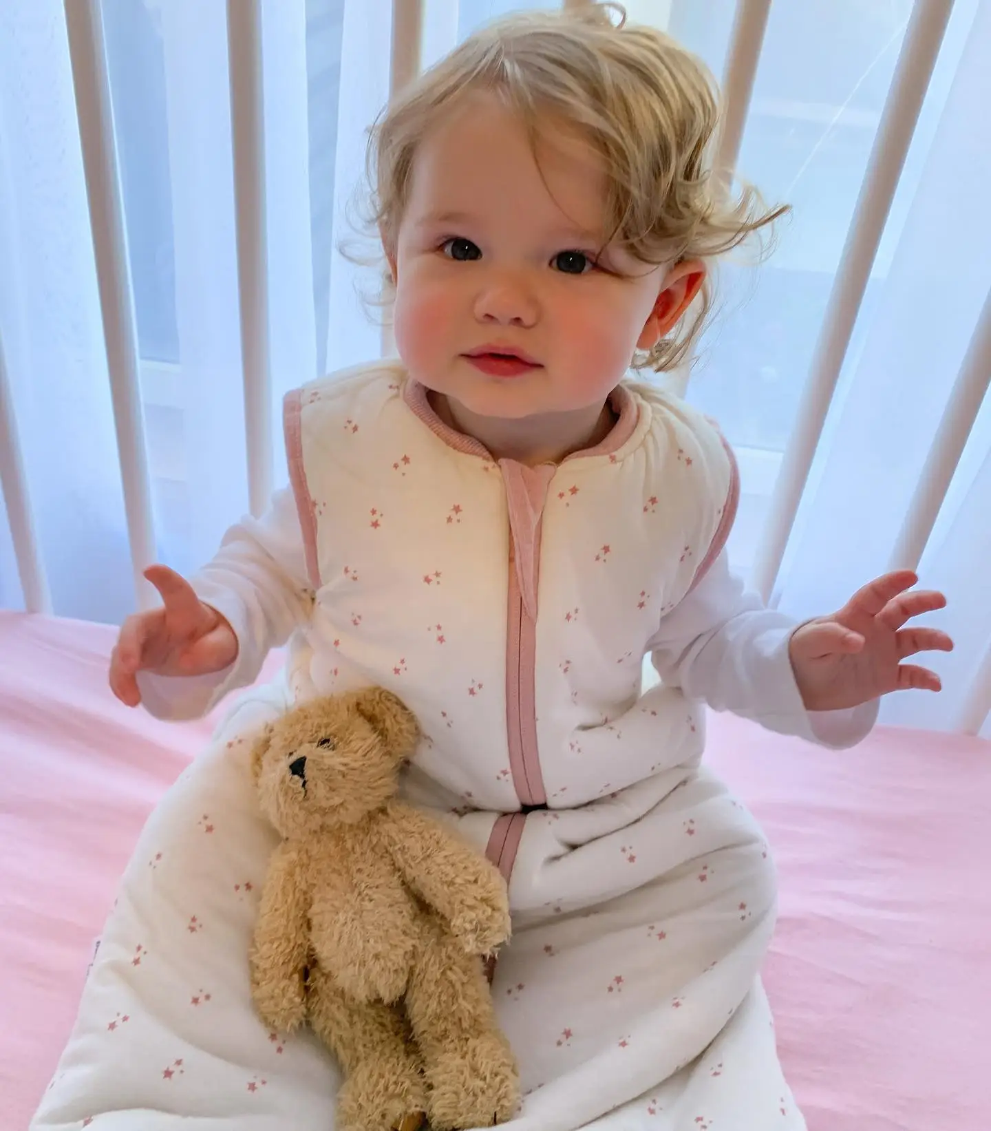 Toddler in a cream star print sleep sack sitting in a crib with a teddy bear