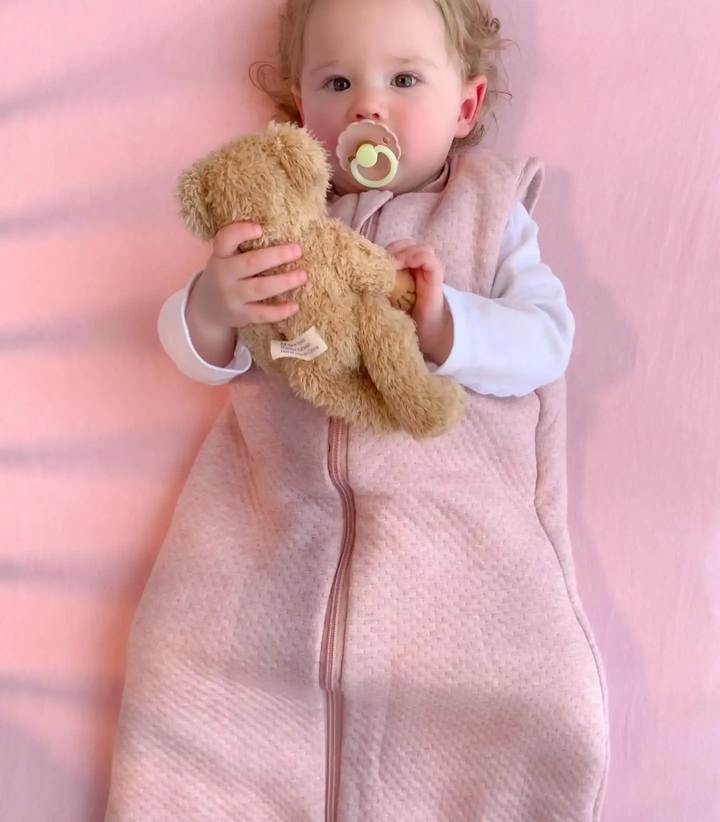 Toddler in a textured pink sleep sack holding a teddy bear on a pink sheet