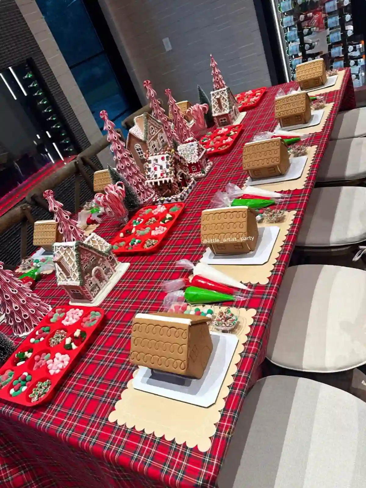 A long table with a red plaid cloth set with gingerbread houses on scalloped placemats, piping bags in red, green, and white, and red muffin trays filled with candy.