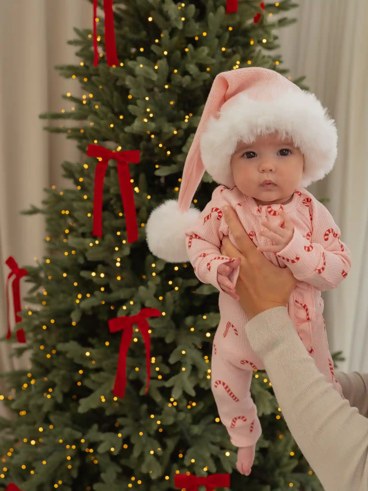 Baby in a long pale pink stocking hat with a white fur band and candy cane waffle knit pajamas in front of a green tree with red velvet ribbons