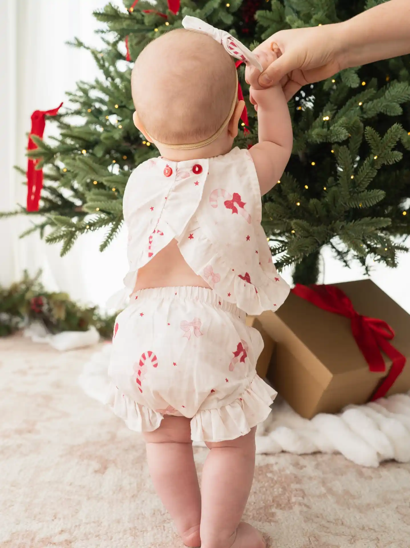 Baby standing from behind in an open back top and ruffled bloomers printed with candy canes and bows beside a gift tied with red velvet ribbon