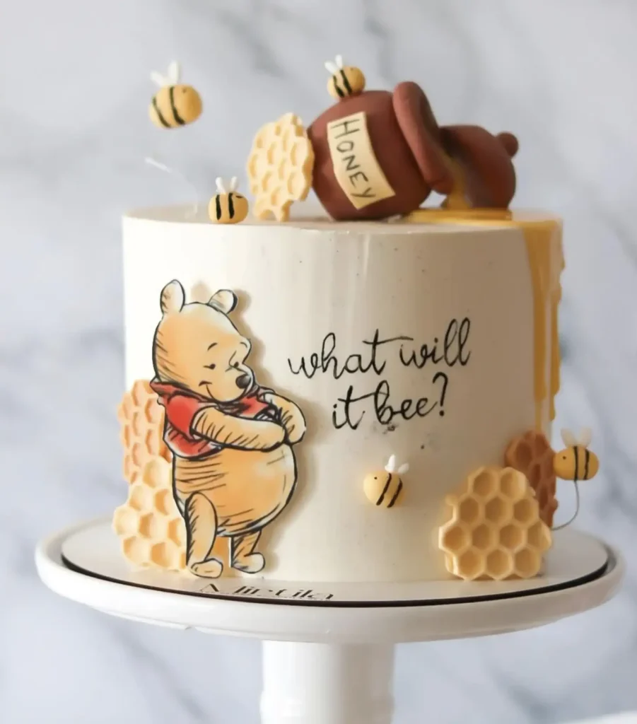 Tall cream cake with a tipped honey pot on top, honey drips, small bees, honeycomb pieces, a Pooh figure, and the words what will it bee