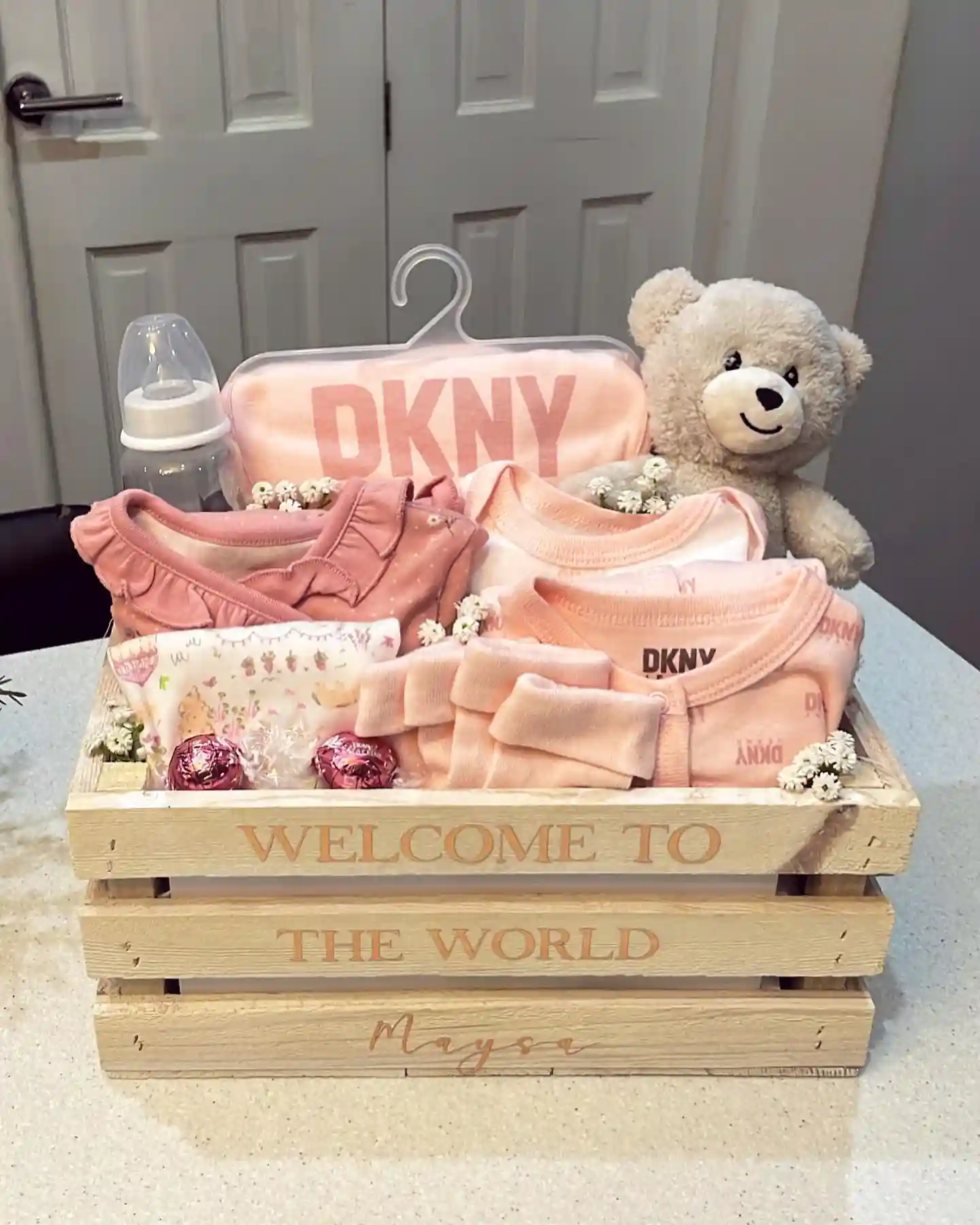 Wooden welcome to the world crate with a stack of soft pink sleepers, a ruffle collar top, a blanket set on a hanger, a baby bottle, a plush bear and two foil wrapped chocolates