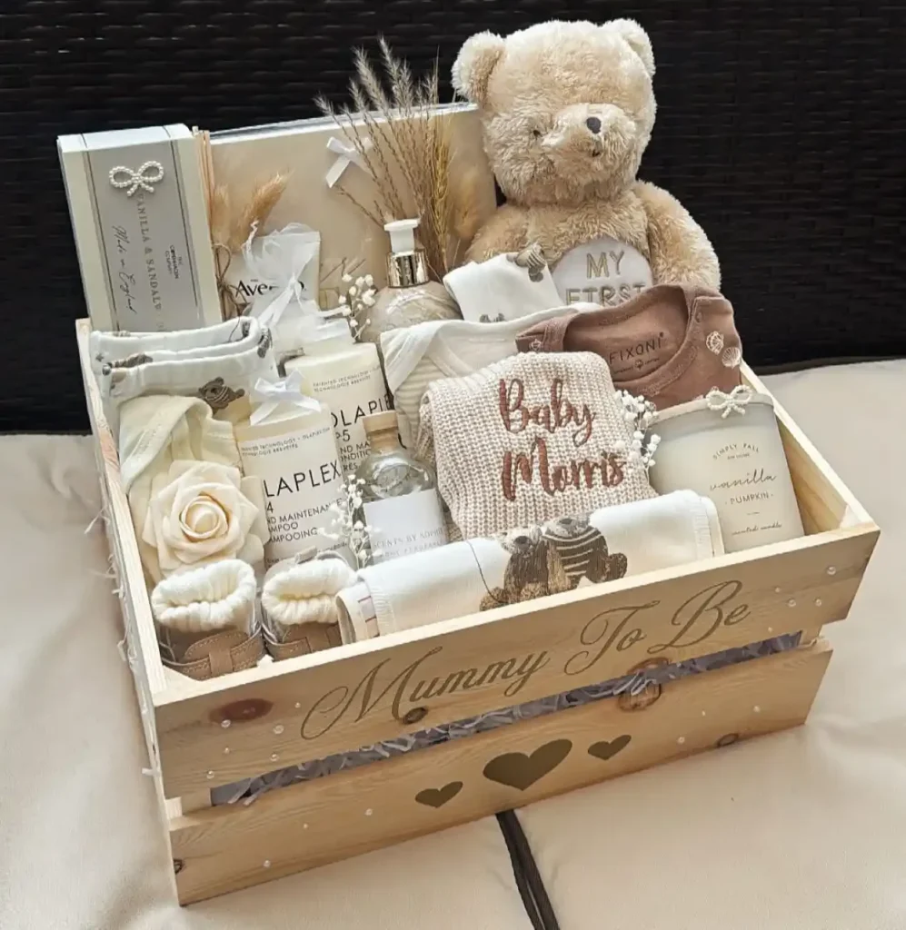 Wooden crate with Mummy To Be in gold script holding a plush bear, an Aveeno tube, hair care bottles, a vanilla pumpkin candle, a reed diffuser, cream knit booties and a knit blanket