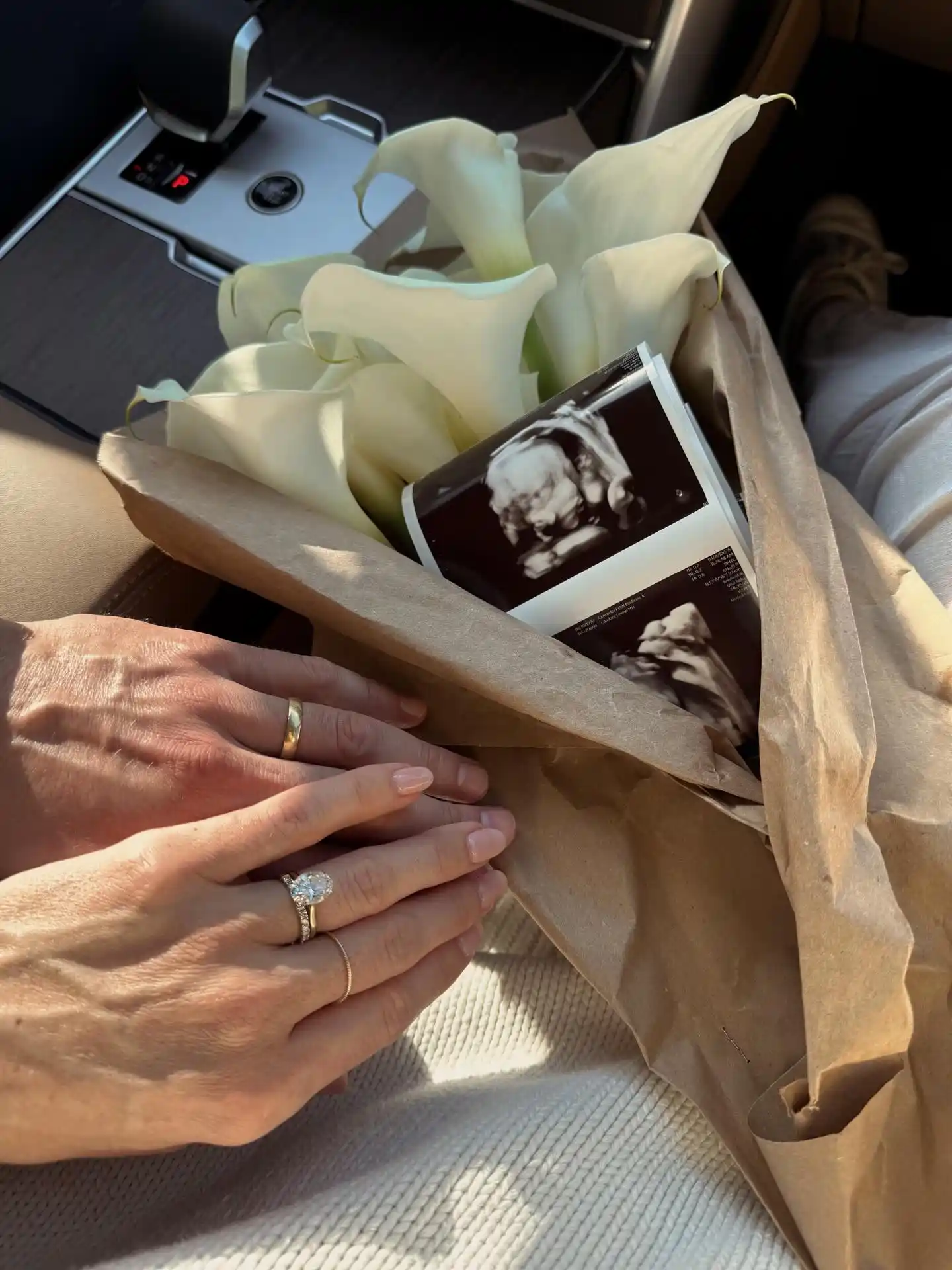 Large white blooms wrapped in brown paper with two scan prints peeking out and a couple's hands resting together on top in warm sun