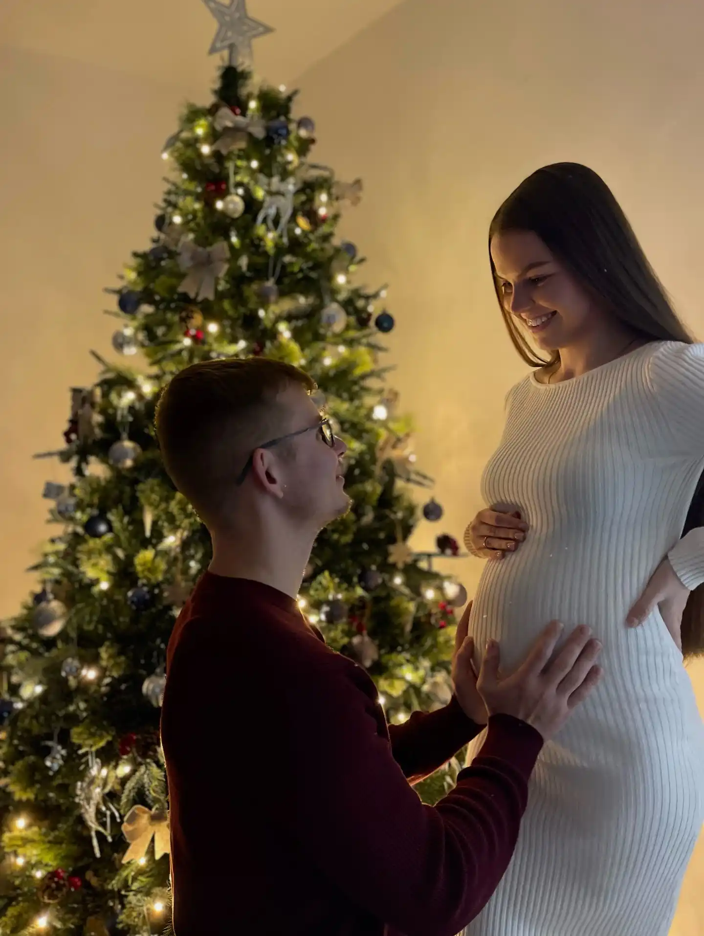 Couple by a lit Christmas tree, the partner kneeling with both hands on the mother-to-be's bump