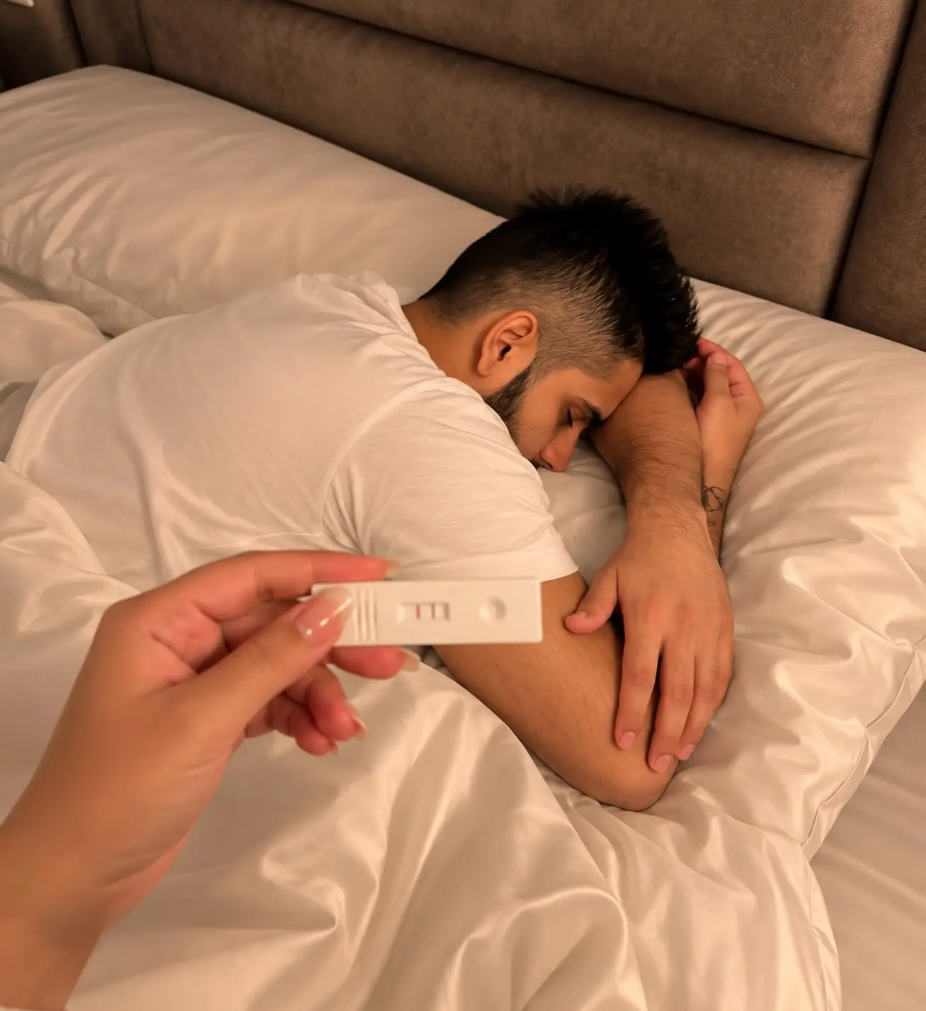 Man sleeping face down on white sheets in front of a brown headboard while a hand holds a pregnancy test over his shoulder, shot from above
