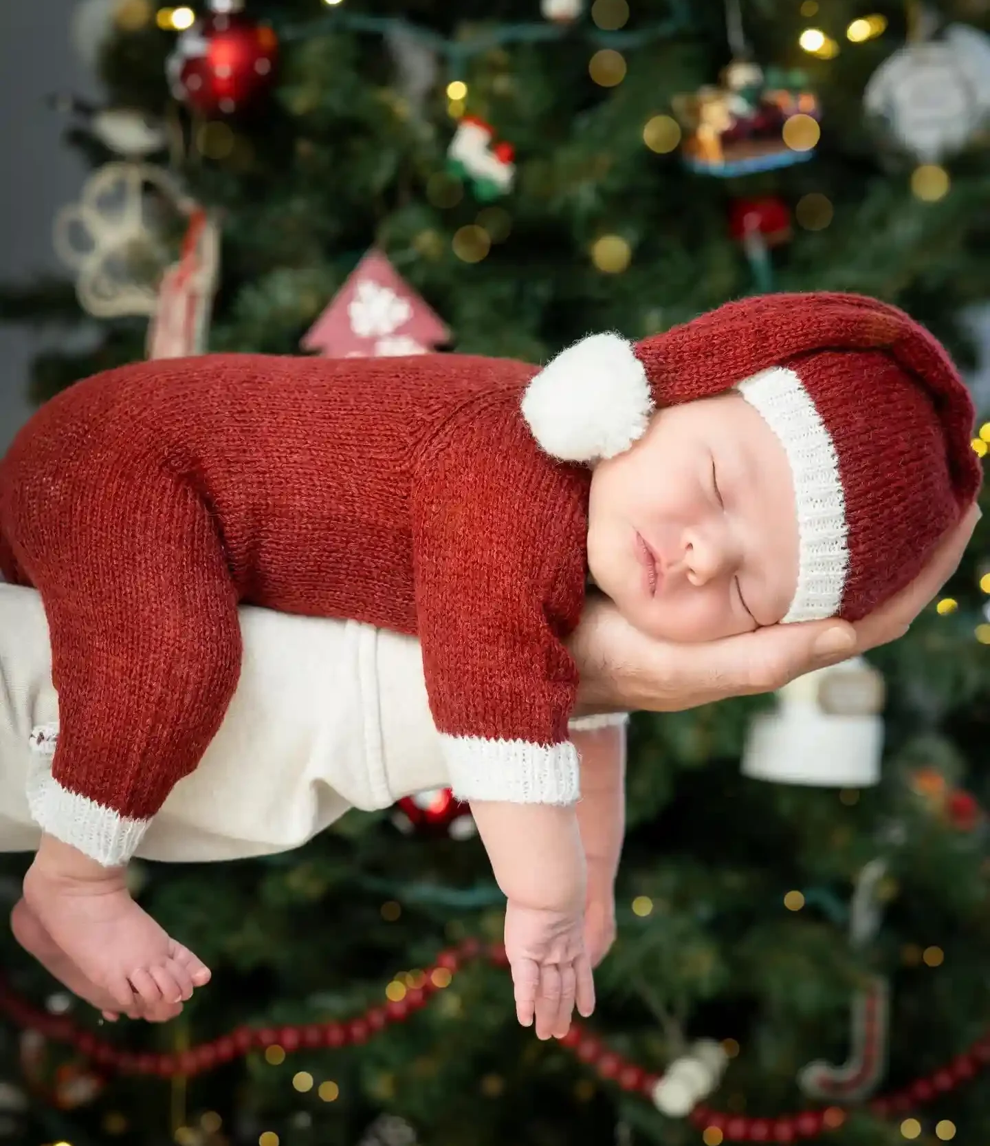 Sleeping newborn resting on a forearm in a red knit romper and long knit hat with a cream pom, with a softly blurred Christmas tree behind