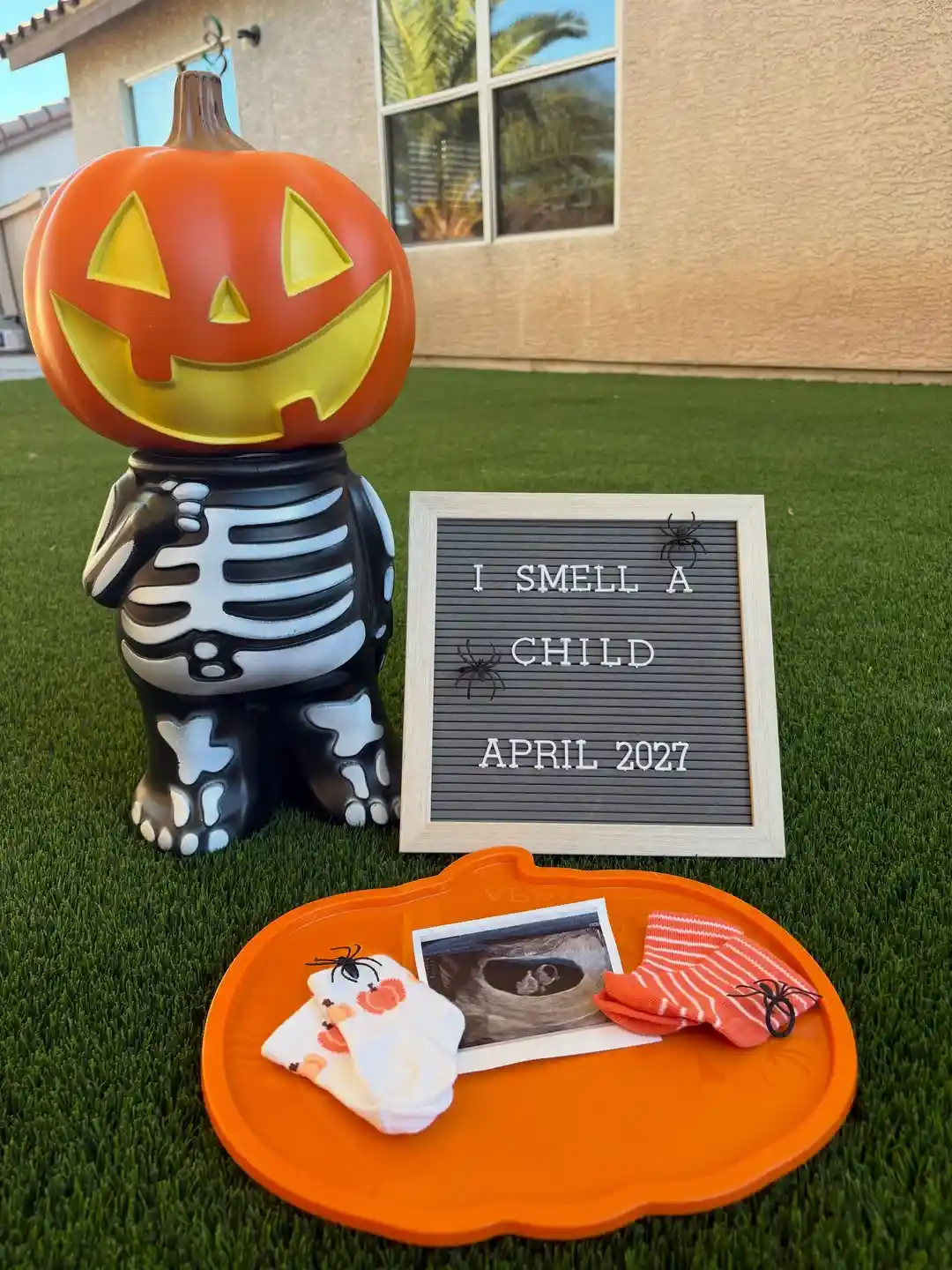 Skeleton figure with a jack-o'-lantern head beside a letter board reading "I smell a child, April 2027" with an ultrasound and baby socks on an orange pumpkin tray on a lawn