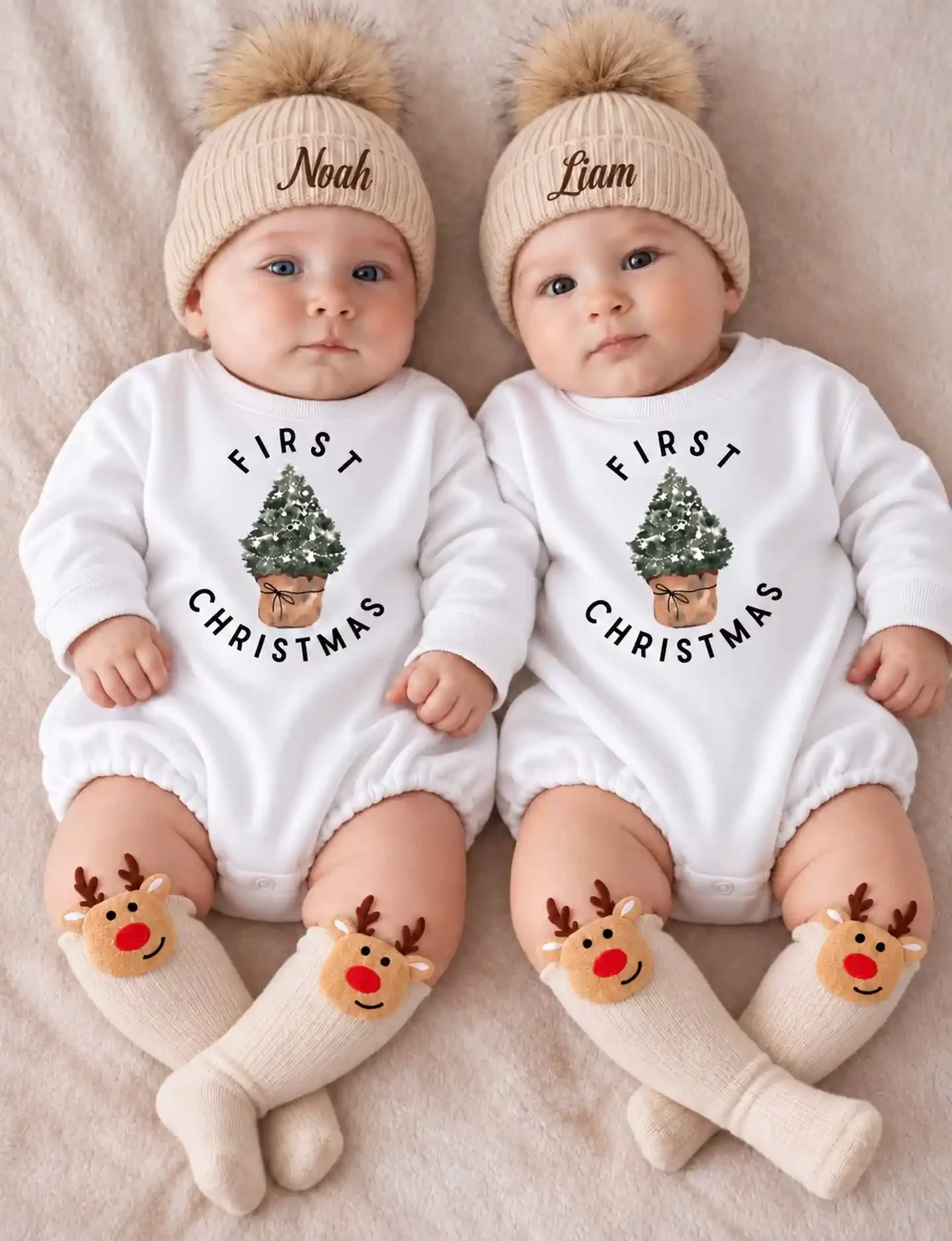Twin babies in matching white First Christmas tree rompers, reindeer knee socks and ribbed beanies with names in brown script, lying side by side on a soft blanket