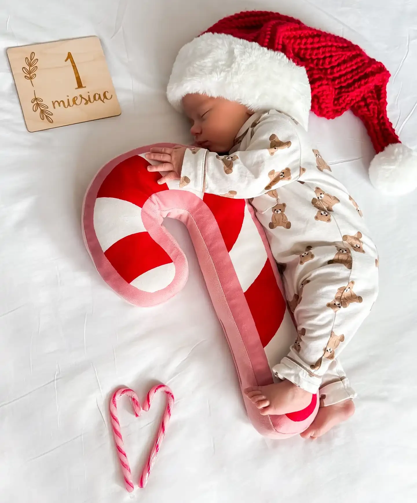 Baby in teddy bear pajamas and a red knit fur band hat curled around a giant candy cane pillow on white sheets beside a wooden one month card
