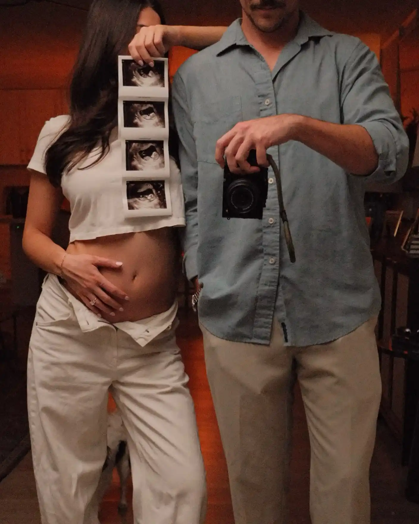 Couple standing side by side in warm dim light, the woman in a white crop top and pants holding a vertical ultrasound strip with a hand on her belly, the man in a blue shirt holding a film camera