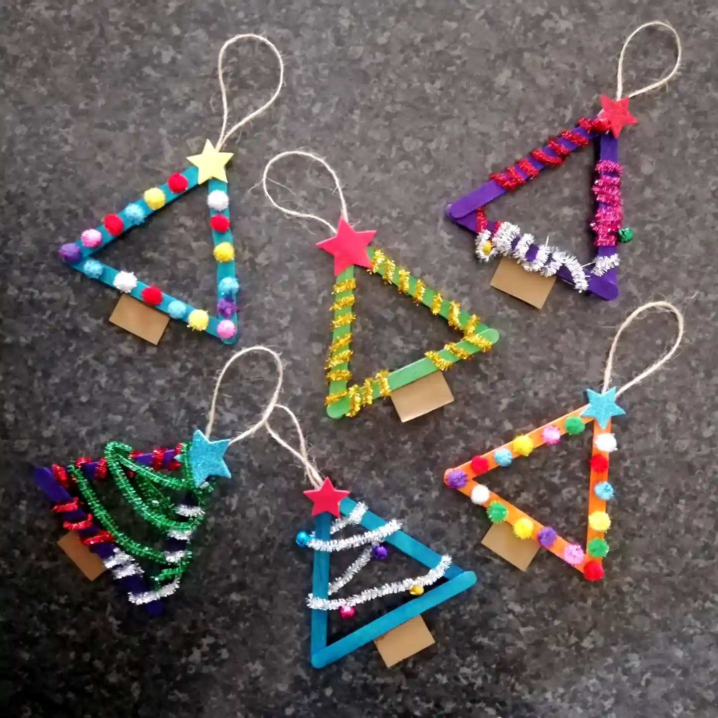 Six triangle shaped popsicle stick Christmas tree ornaments in bright colors, decorated with pom poms, pipe cleaners, foam stars, and small bells, each with a twine loop.