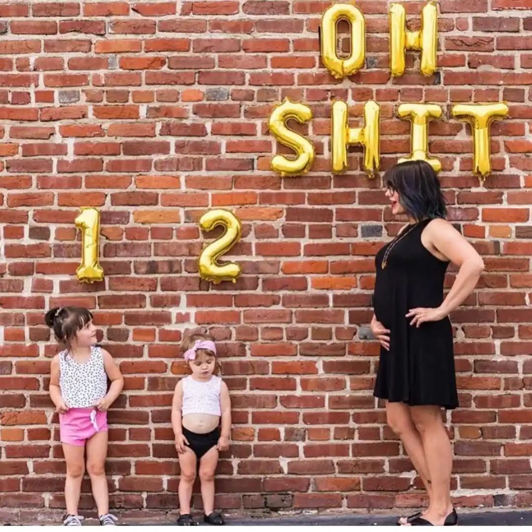 Pregnant woman in a black dress facing two toddlers against a red brick wall, with gold foil balloons spelling "Oh sh*t" and the numbers 1 and 2