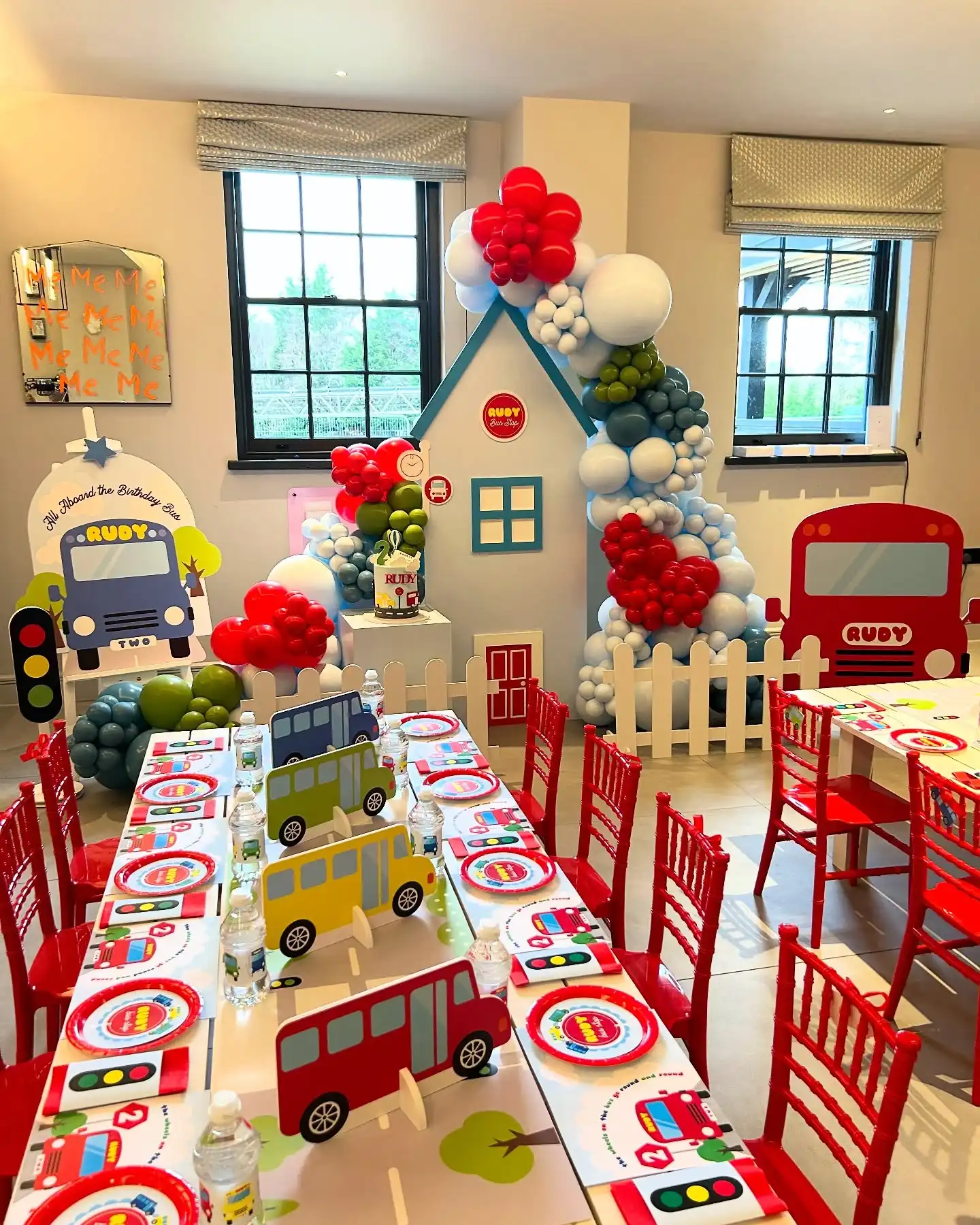 Bus birthday table with a paper road down the middle, trees and flat red, yellow, green and navy bus cutouts, bus and traffic light plates, red child size chairs, a bus stop house backdrop with a white picket fence and an All Aboard sign on an easel