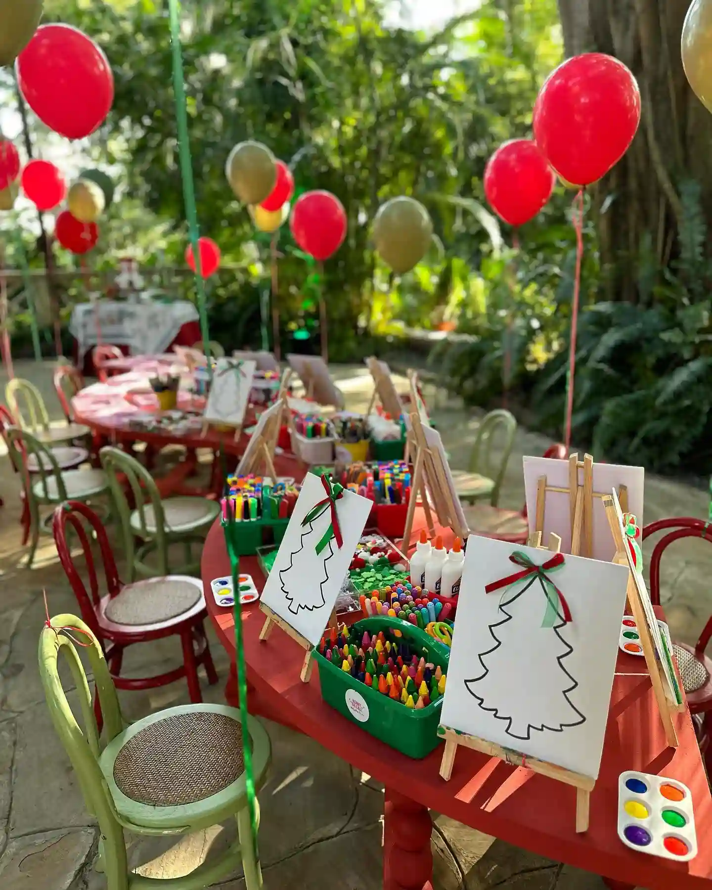 A red children's table outdoors with mini easels holding canvases with a Christmas tree outline and ribbon bow, crayon and marker caddies, glue, and paint palettes, with red and green balloons behind.