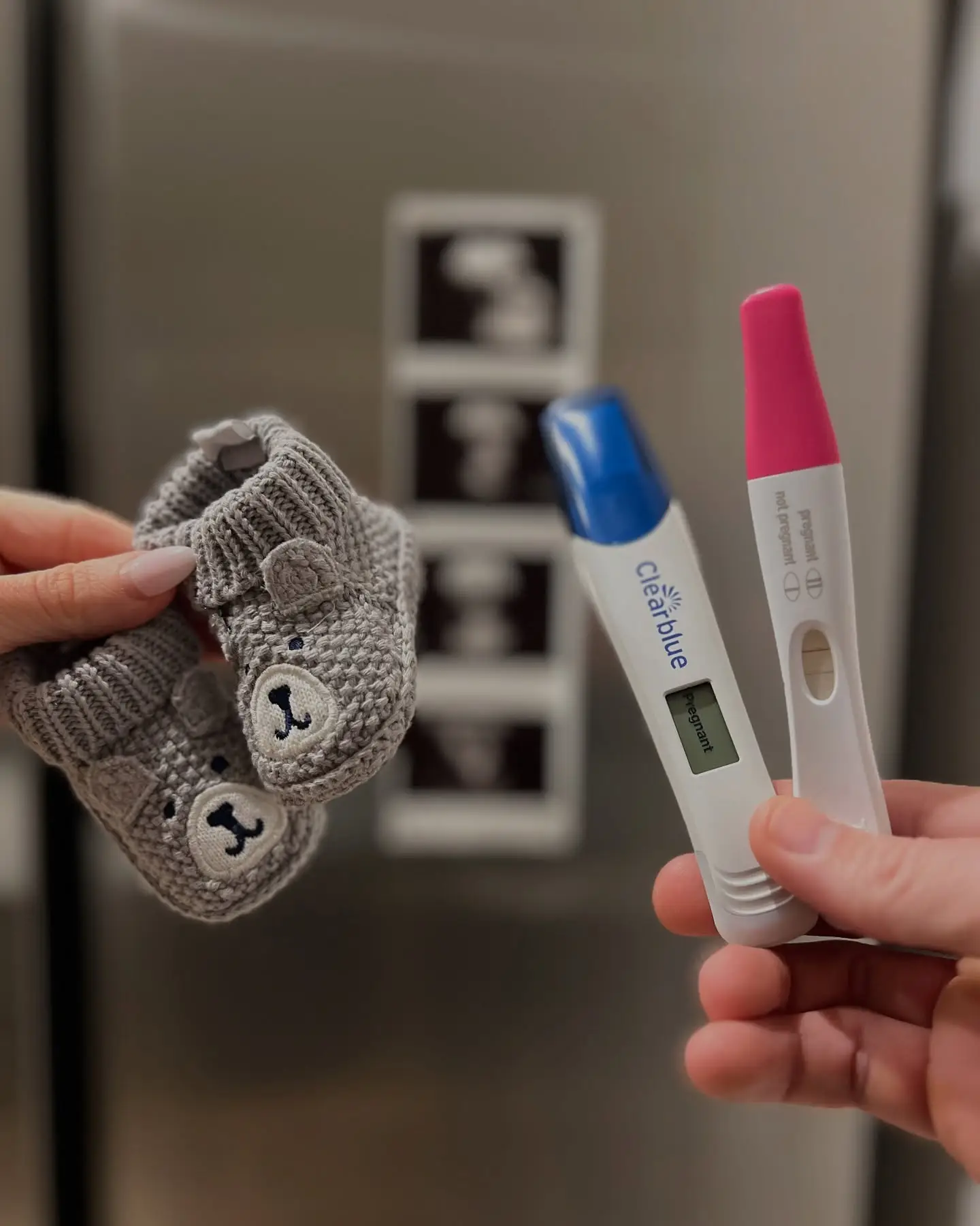Two hands holding gray knit bear booties and two pregnancy tests in front of a steel fridge covered with small scan photos