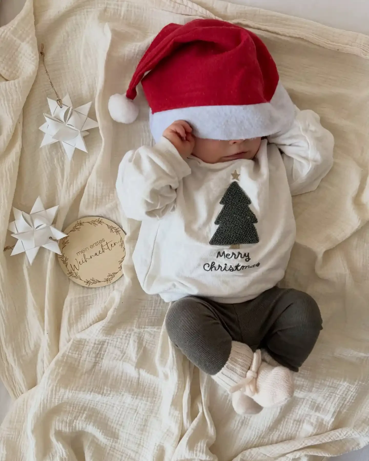 Baby in a cream sweatshirt with a fuzzy green tree and Santa hat beside a round wooden my first Christmas plaque and folded paper stars on a muslin blanket