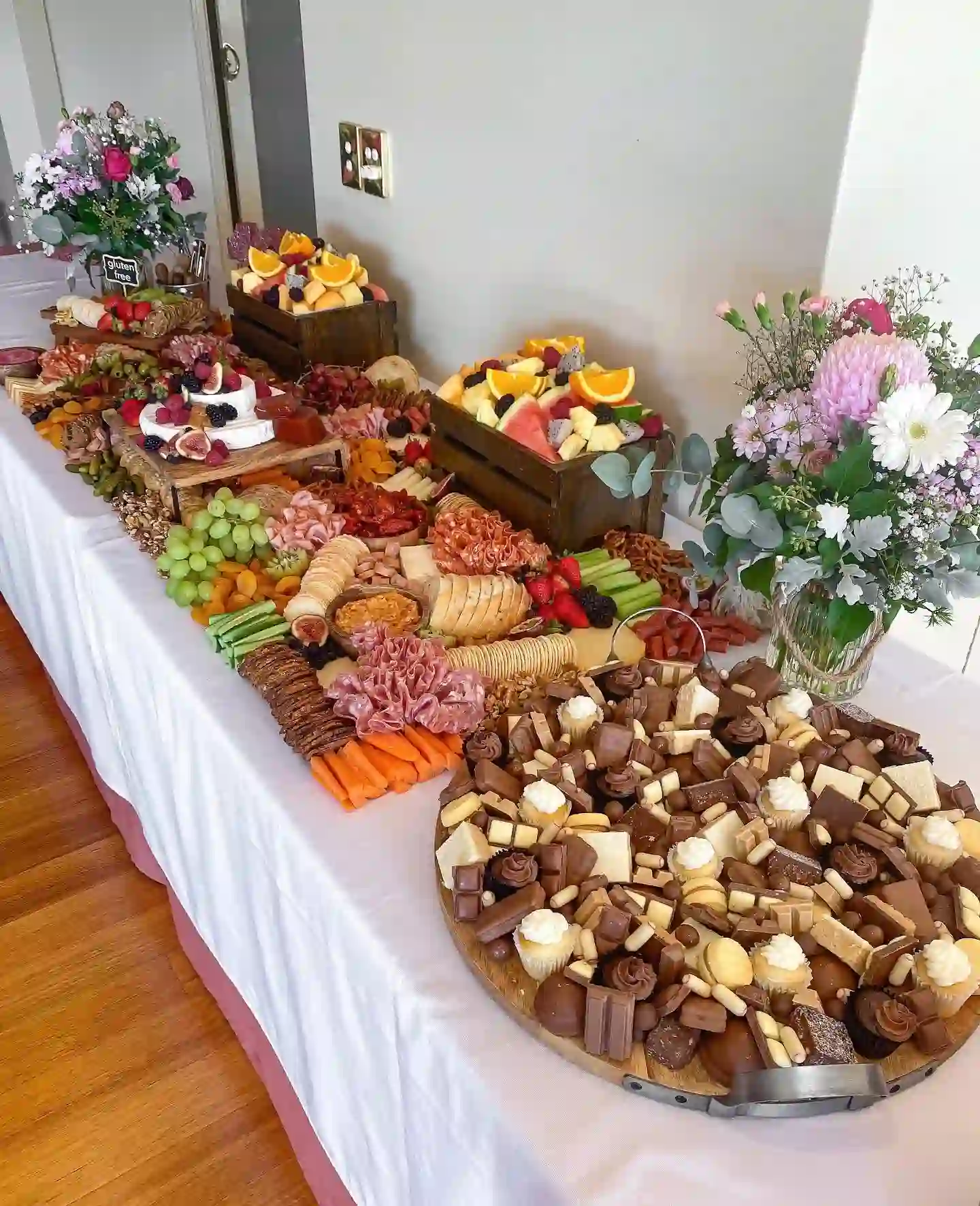 Long white grazing table with cheese, cured meats, crackers, fruit crates, a gluten free sign and a big round tray of chocolates and cupcakes