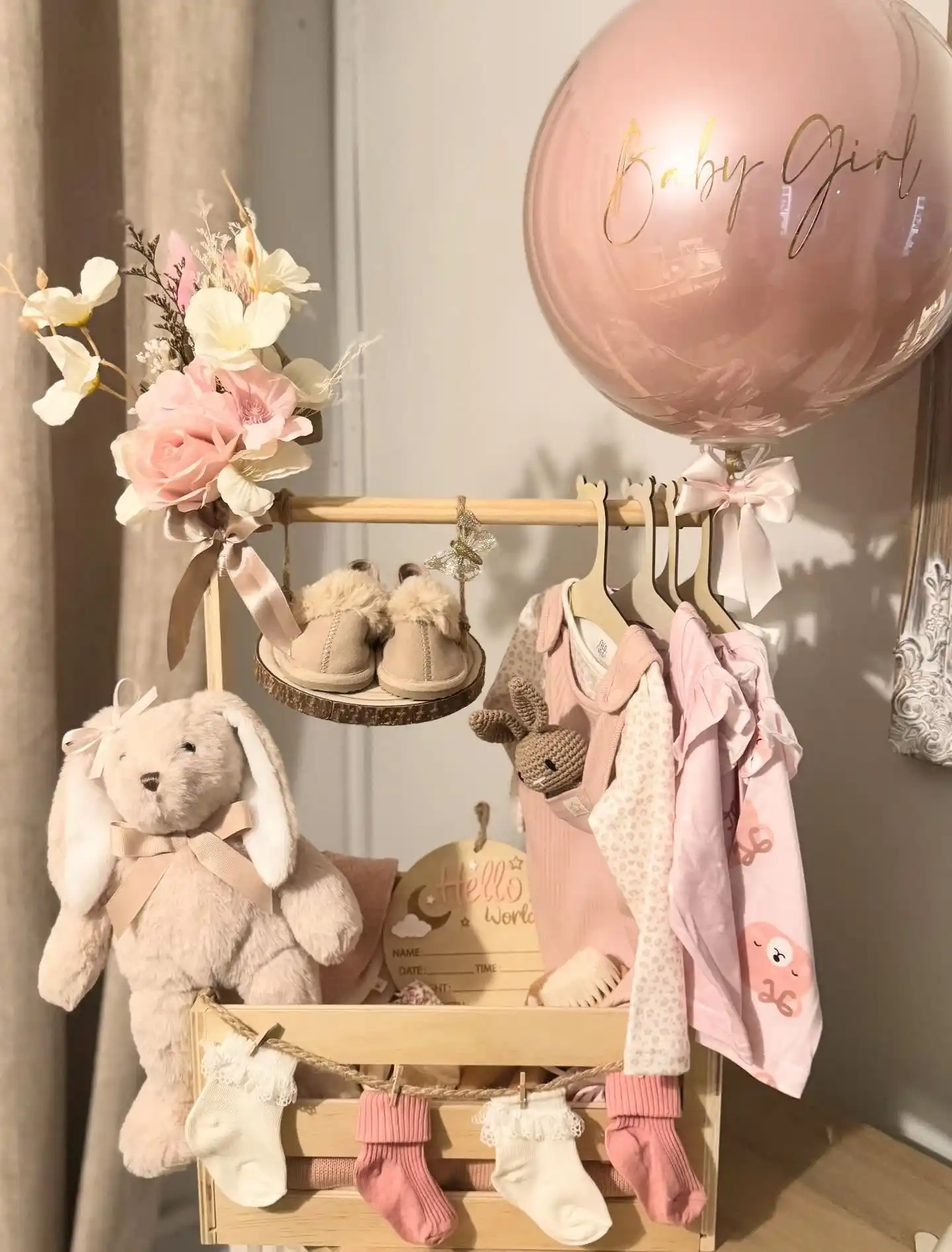 Small wooden clothes rail with pink and cream baby outfits on tiny hangers, socks pegged to a twine garland, a plush bunny, fuzzy booties, a Hello World plaque and a blush balloon