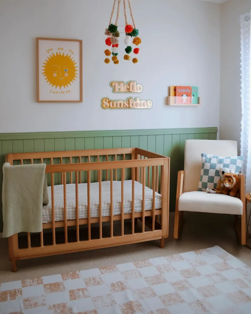 Nursery with green slatted paneling, a natural wood crib, a sun print, a Hello Sunshine sign and a pom pom mobile