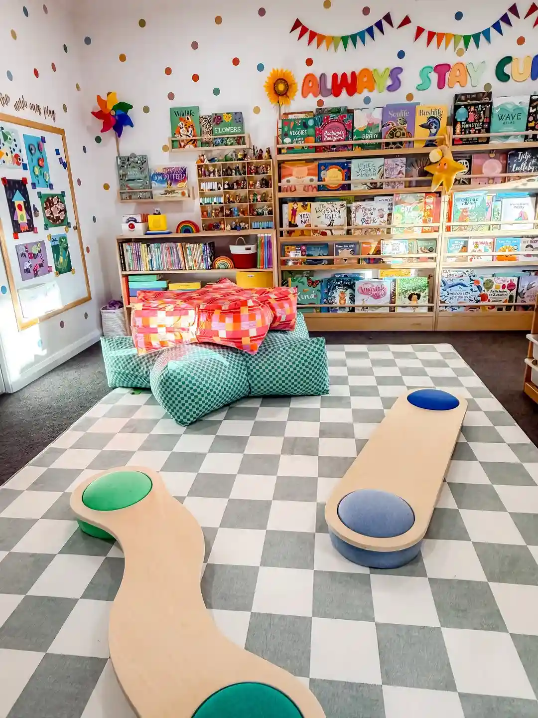 Playroom wall with narrow ledges showing books cover out above a checkered rug, a pile of floor cushions and two curvy balance boards
