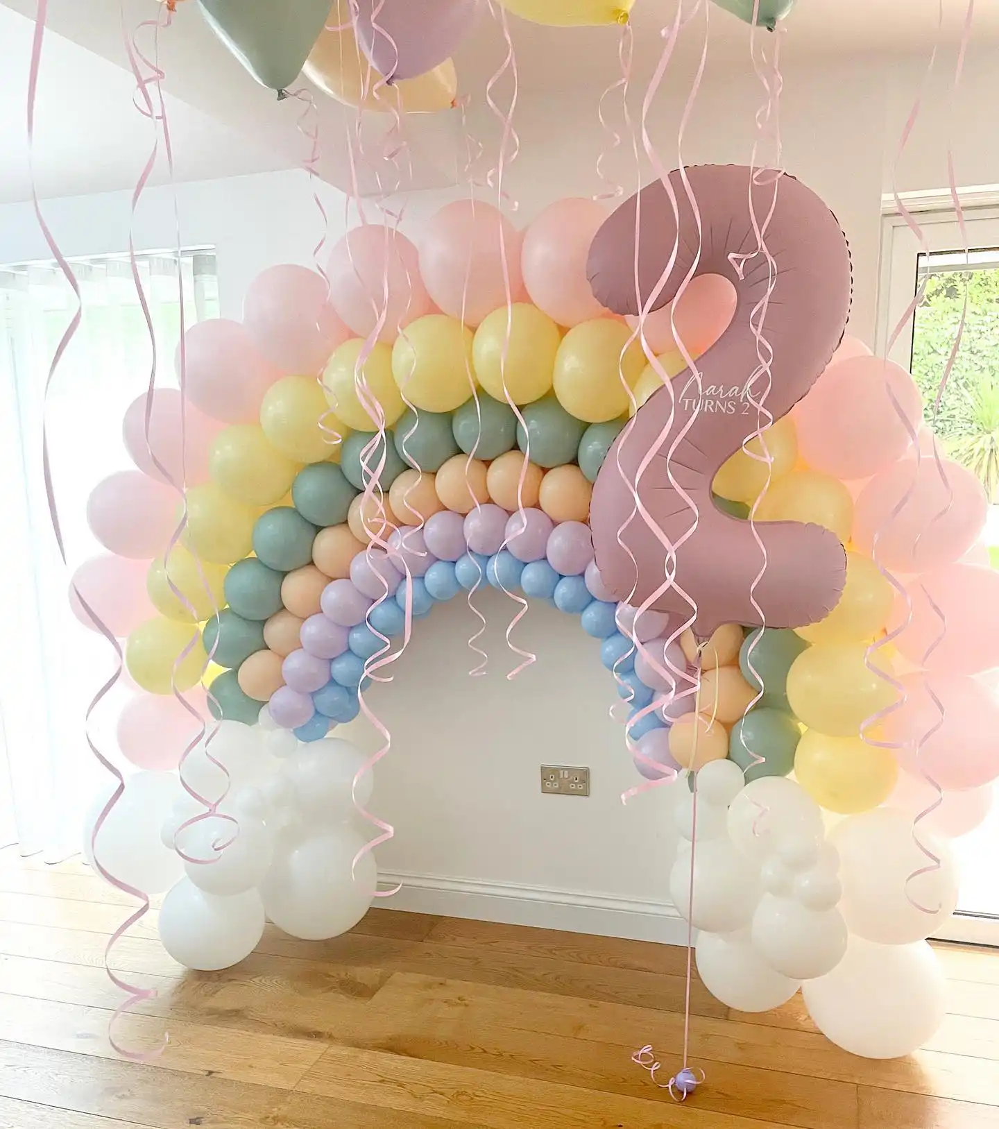 Pastel rainbow balloon arch on a white wall in blue, lilac, peach, sage green, yellow and soft pink stripes with white balloon clouds, a mauve foil number 2 and curly pink ribbons