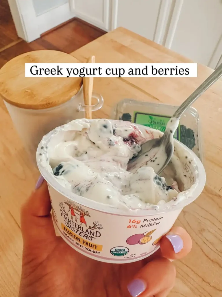 Hand holding a passion fruit skyr yogurt cup stirred with blackberries and chia seeds, with a spoon resting in the cup