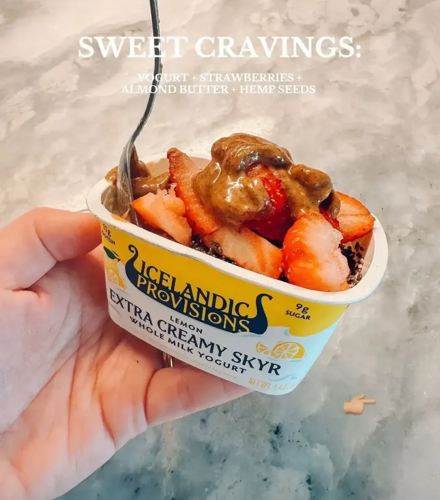 Hand holding a lemon skyr yogurt cup topped with sliced strawberries, almond butter and hemp seeds, with a spoon in the cup