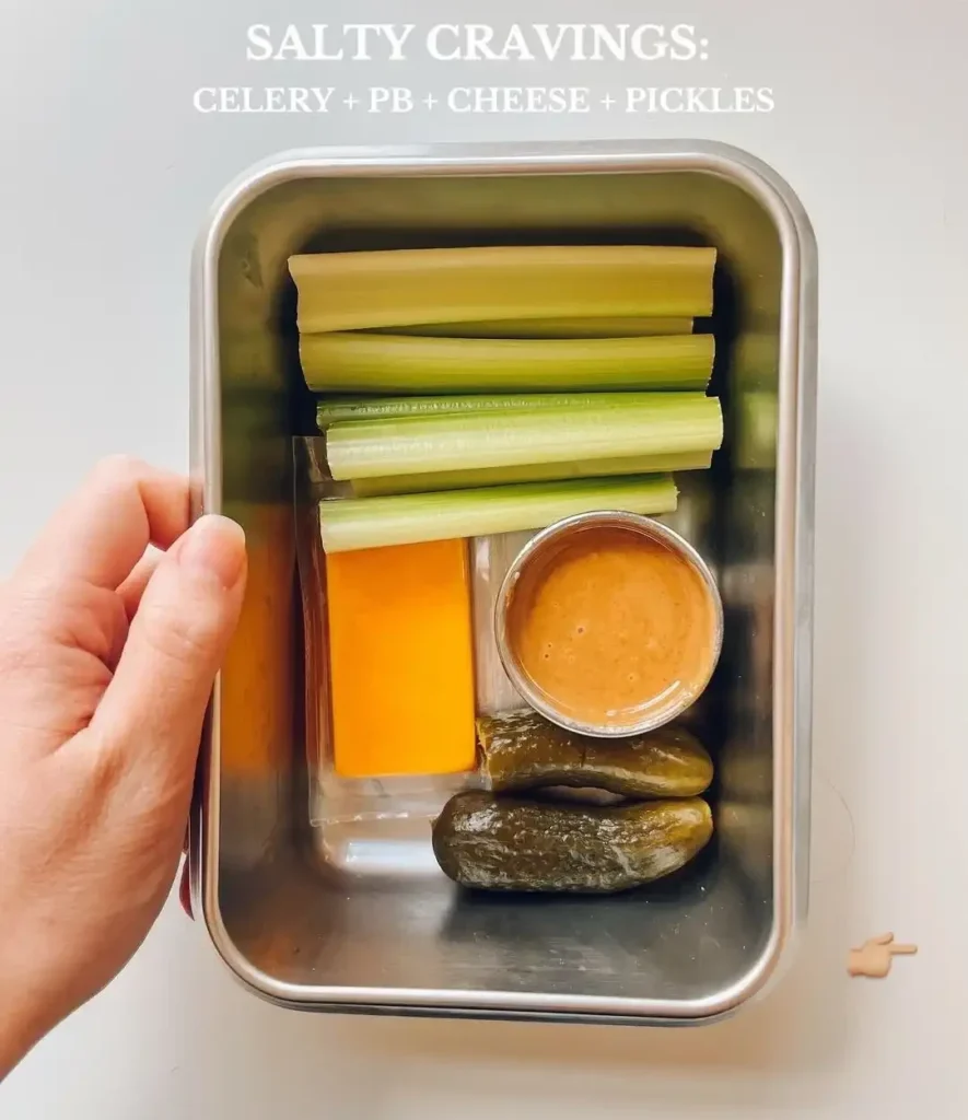 Metal lunch box holding celery sticks, a block of cheddar cheese, a small cup of peanut butter and two pickles