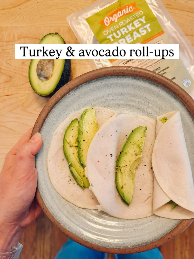 Plate with slices of oven roasted turkey breast topped with avocado slices and black pepper, one slice already rolled up