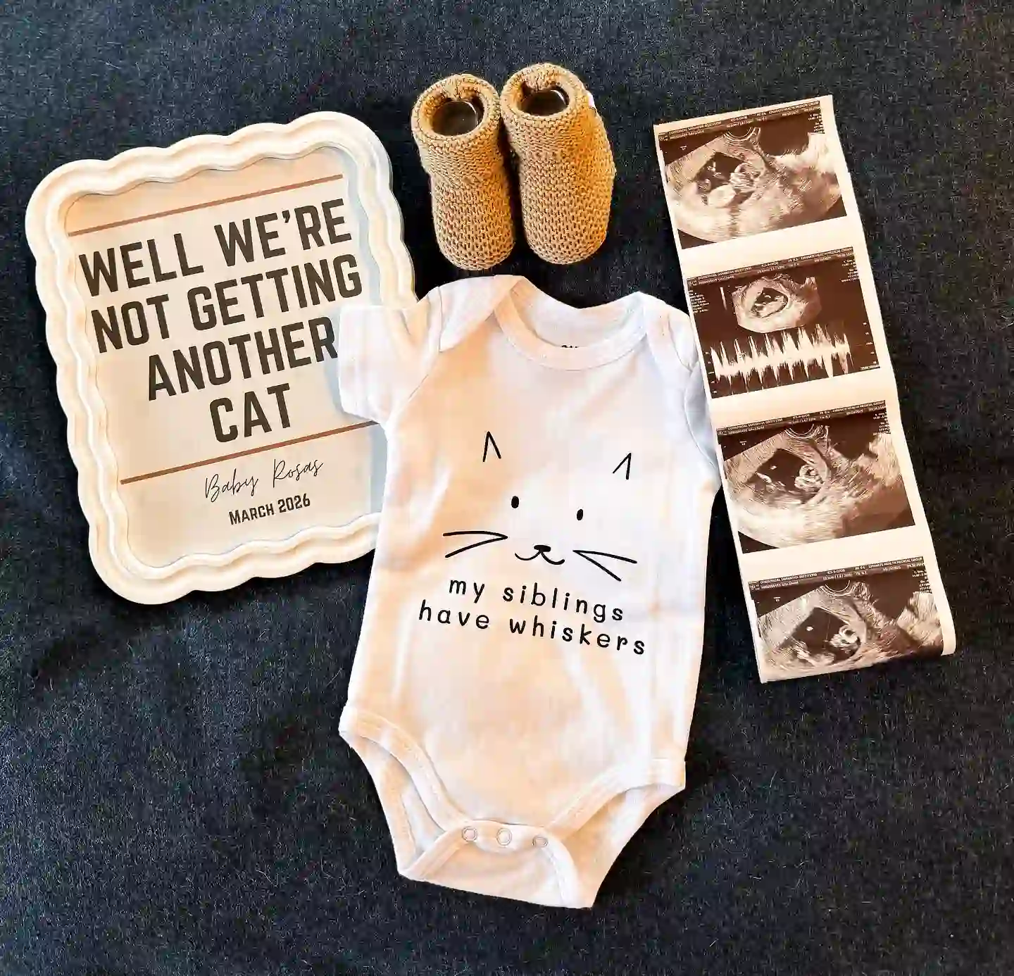 Flat lay on dark fabric with a wavy frame reading "Well we're not getting another cat", a white onesie with a cat face reading "my siblings have whiskers", knit booties and an ultrasound strip