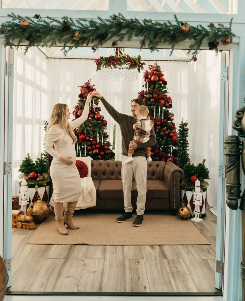 Pregnant mom twirling beside dad holding a toddler in front of red Christmas trees for a family holiday card