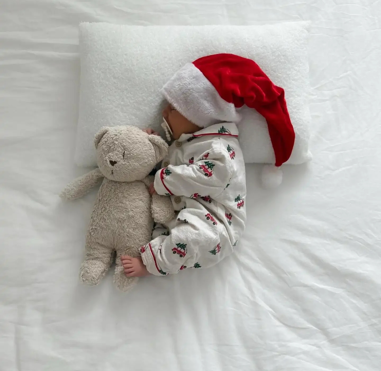 Baby in flannel pajamas printed with red cars carrying trees, a plush Santa hat and a beige teddy on white sheets, seen from above