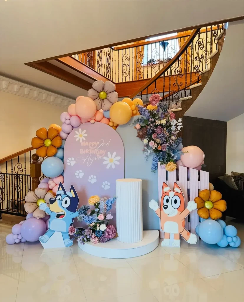 Bluey and Bingo cutouts on a lilac arch with white daisies and paw prints, tan and rust foil daisy balloons, flower arrangements in blue, pink and purple, a pink picket fence panel and a white pillar