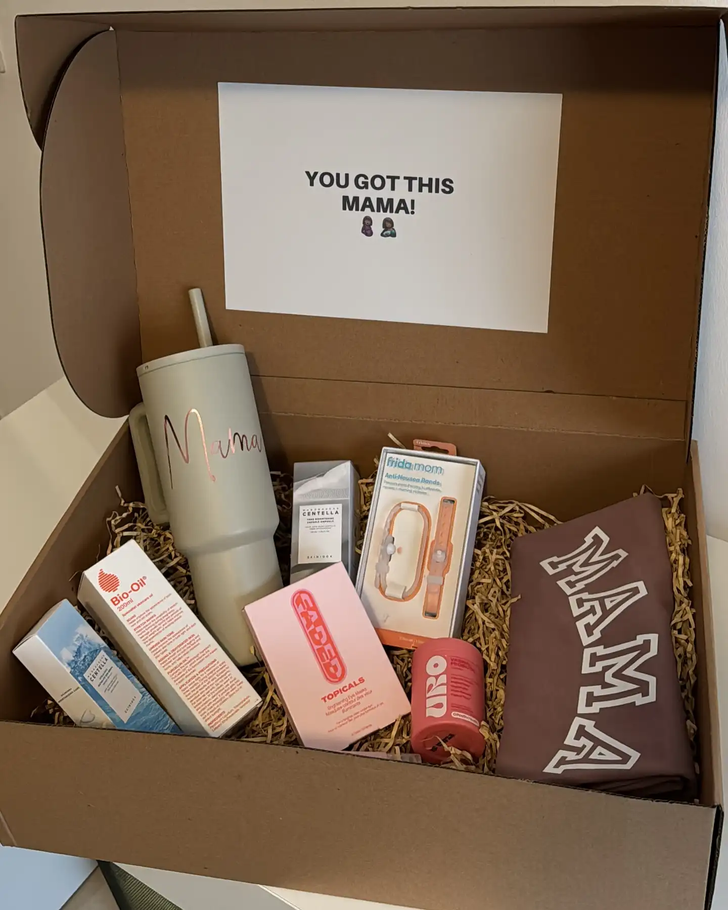 Kraft gift box for a new mom with a big straw tumbler with her name, Bio-Oil, face care, brightening eye masks, a brown MAMA shirt and a card reading You got this mama