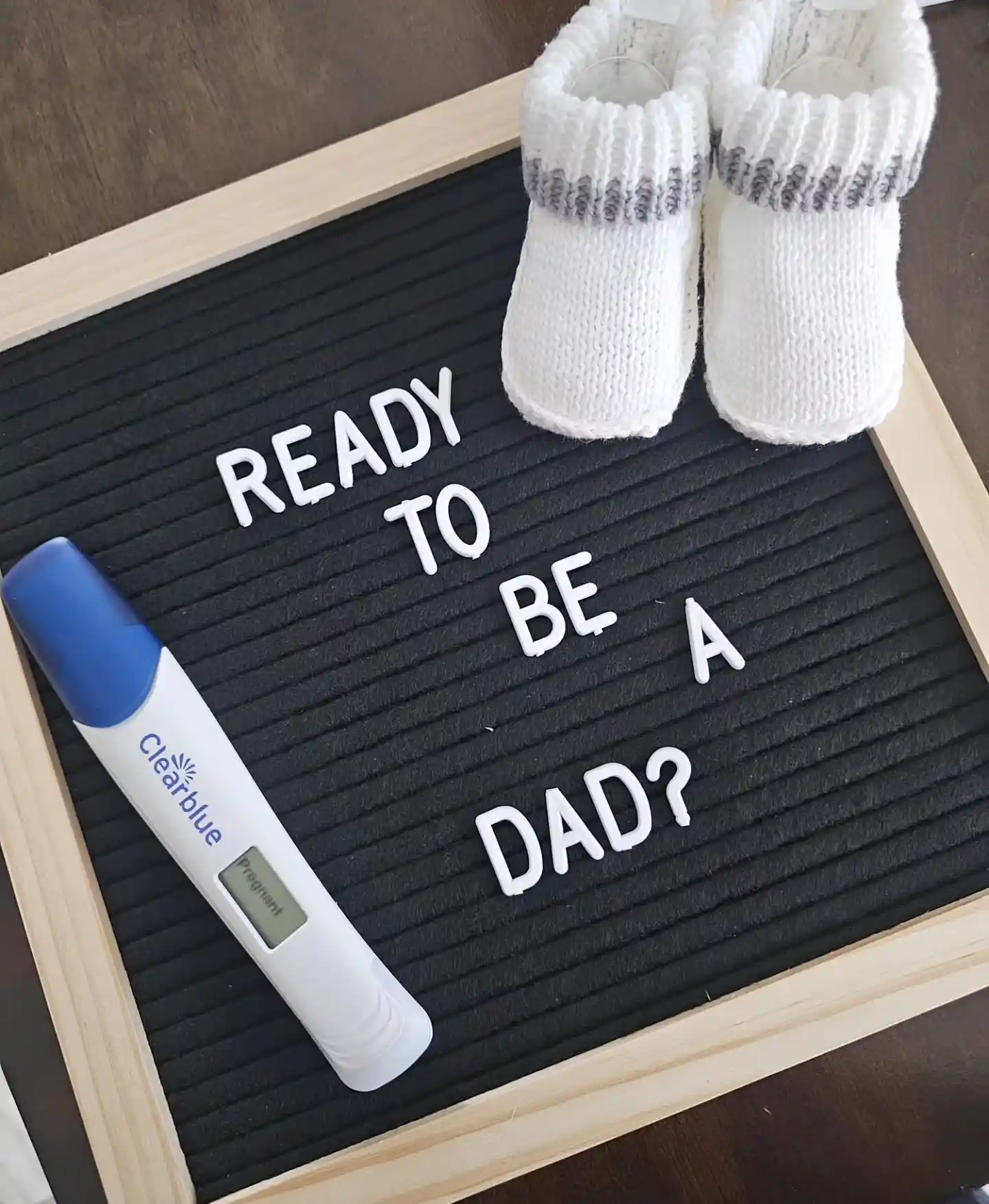 Black letter board reading "Ready to be a dad?" with a digital pregnancy test showing Pregnant and a pair of white knit baby booties