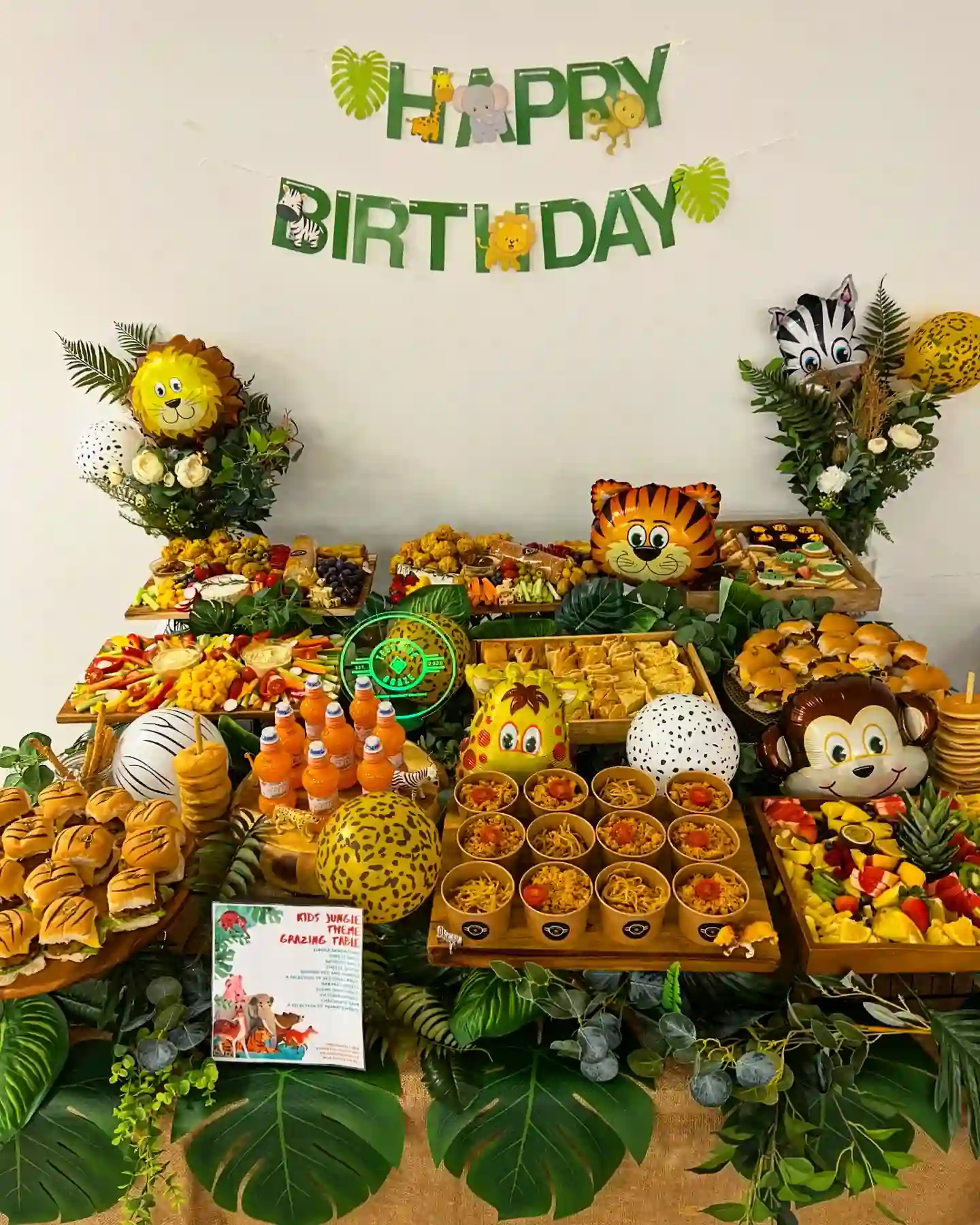 Jungle birthday grazing table with tiger-striped sliders, cups of noodles, fruit tray, veggie sticks and animal balloons