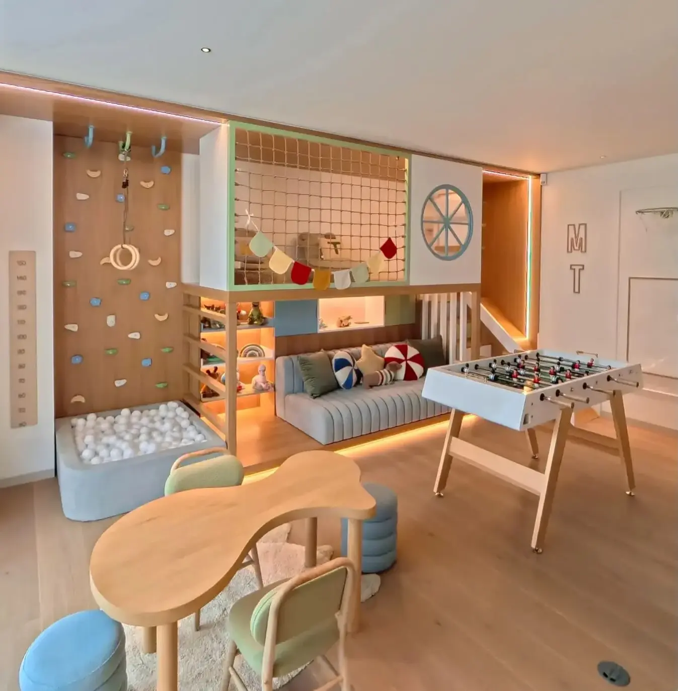 Playroom with a raised net front loft above a low sofa and lit open shelves, a climbing wall, a ball pit, a foosball table and a curvy table with two chairs