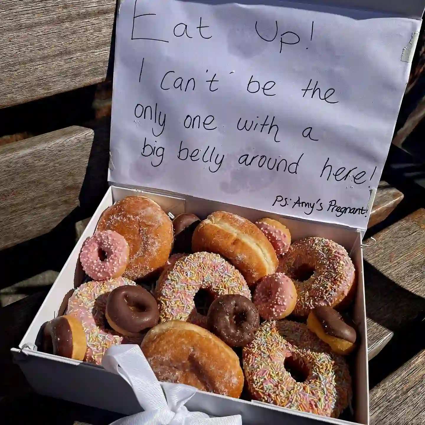 Open white box of sprinkle, chocolate and sugar donuts with a handwritten note inside the lid reading "Eat up! I can't be the only one with a big belly around here! PS: Amy's pregnant!"