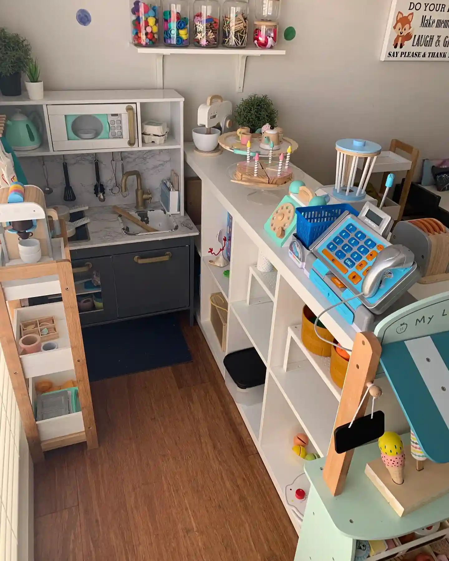 White cube shelf used as a shop counter with a cash register, toy cake and wooden blender on top, baskets in the cubbies, jars of pom poms and a small wooden cart beside a play kitchen