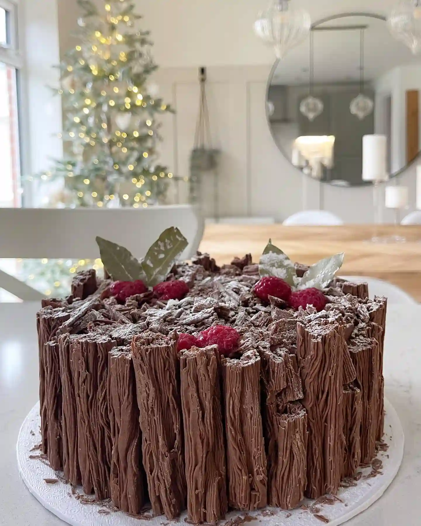 Round cake lined with upright chocolate flake bars and topped with crushed flake, raspberries and sugar dusted bay leaves with a lit tree behind