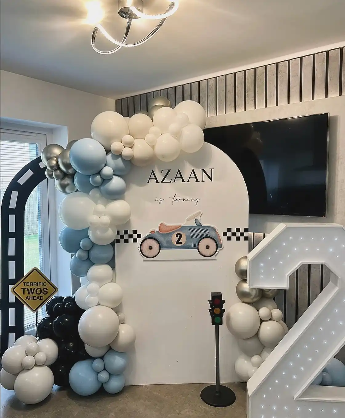 White arch with a small vintage race car with a 2 on its door and a checkered strip, powder blue, cream, navy and silver balloons, a toy traffic light, a yellow Terrific Twos Ahead sign and a tall white number 2 covered in tiny lights