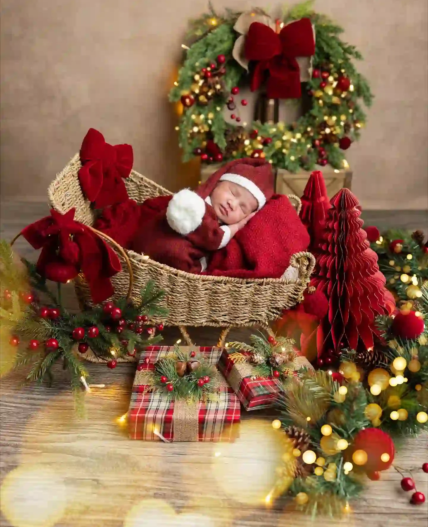Sleeping newborn in a red knit suit and long hat inside a woven sleigh basket with a velvet bow, wreath, honeycomb paper trees and plaid wrapped gifts