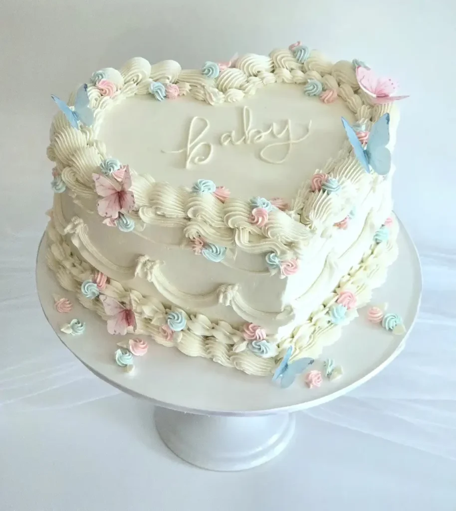 Cream heart shaped cake with the word baby piped in script, wavy piped swags, pink and blue rosettes, and pink and blue butterflies