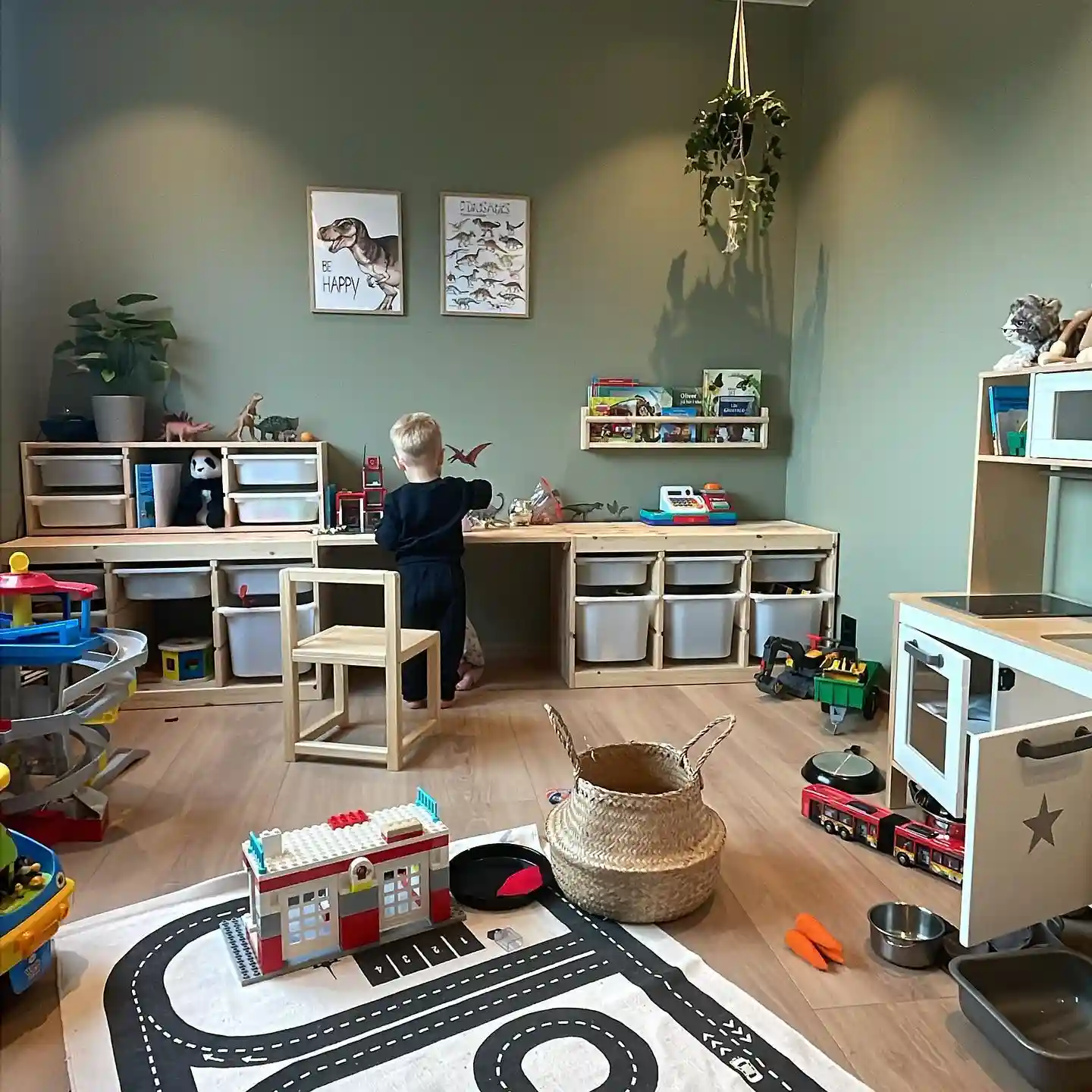 Playroom with a sage green wall, a long wooden frame of white tubs topped with a play counter, a book ledge, a woven basket and a road rug with a fire station build and toy bus