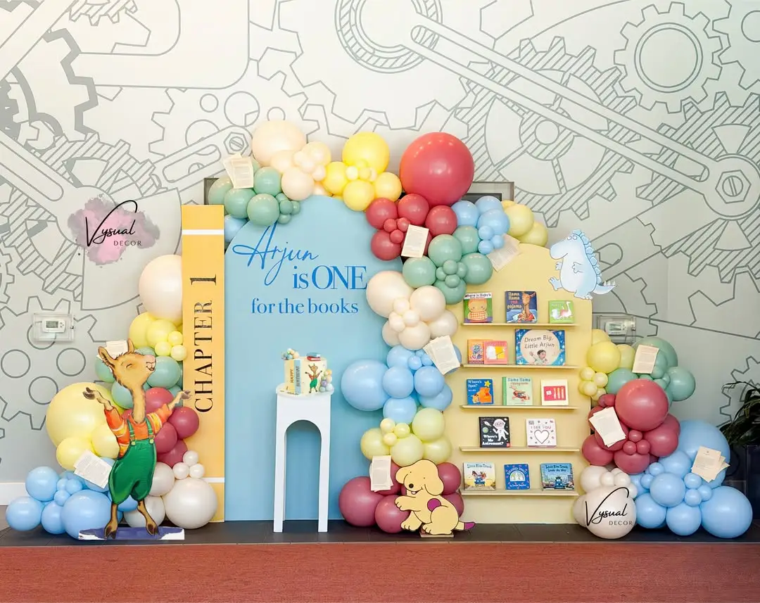 Storybook first birthday setup with a pastel balloon garland, a blue arch backdrop and a shelf of picture books